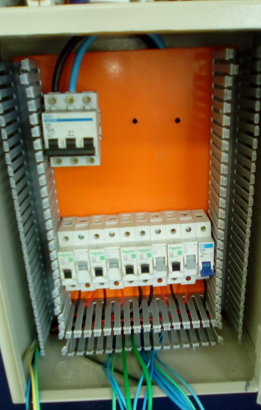 quadro de distribuição elétrica interno com disjuntores, fiação colorida e canaletas, fundo laranja  — texto:  