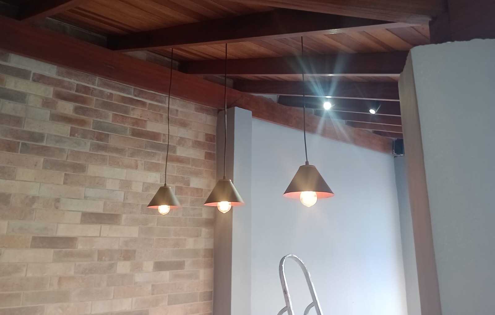 três pendentes cone de metal acesos pendurados do teto de madeira sobre parede de tijolinho aparente, com uma 